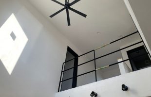 吹き抜けのあるリビング空間。白い塗り壁に自然光が差し込み、黒いシーリングファンとアイアン手すりがアクセントになった新築住宅の内観。