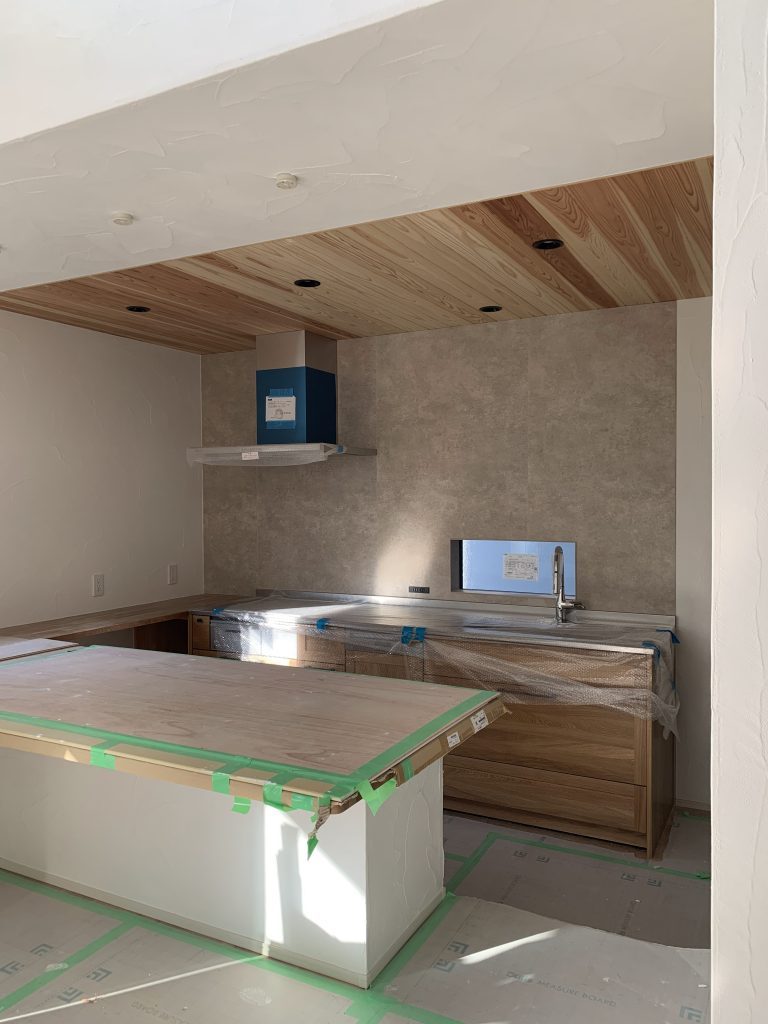 杉板の木天井と木目のキッチン収納が特徴のナチュラルモダンな新築キッチン。ニュアンスグレーの壁と大きなカウンターが設置された施工中の様子。